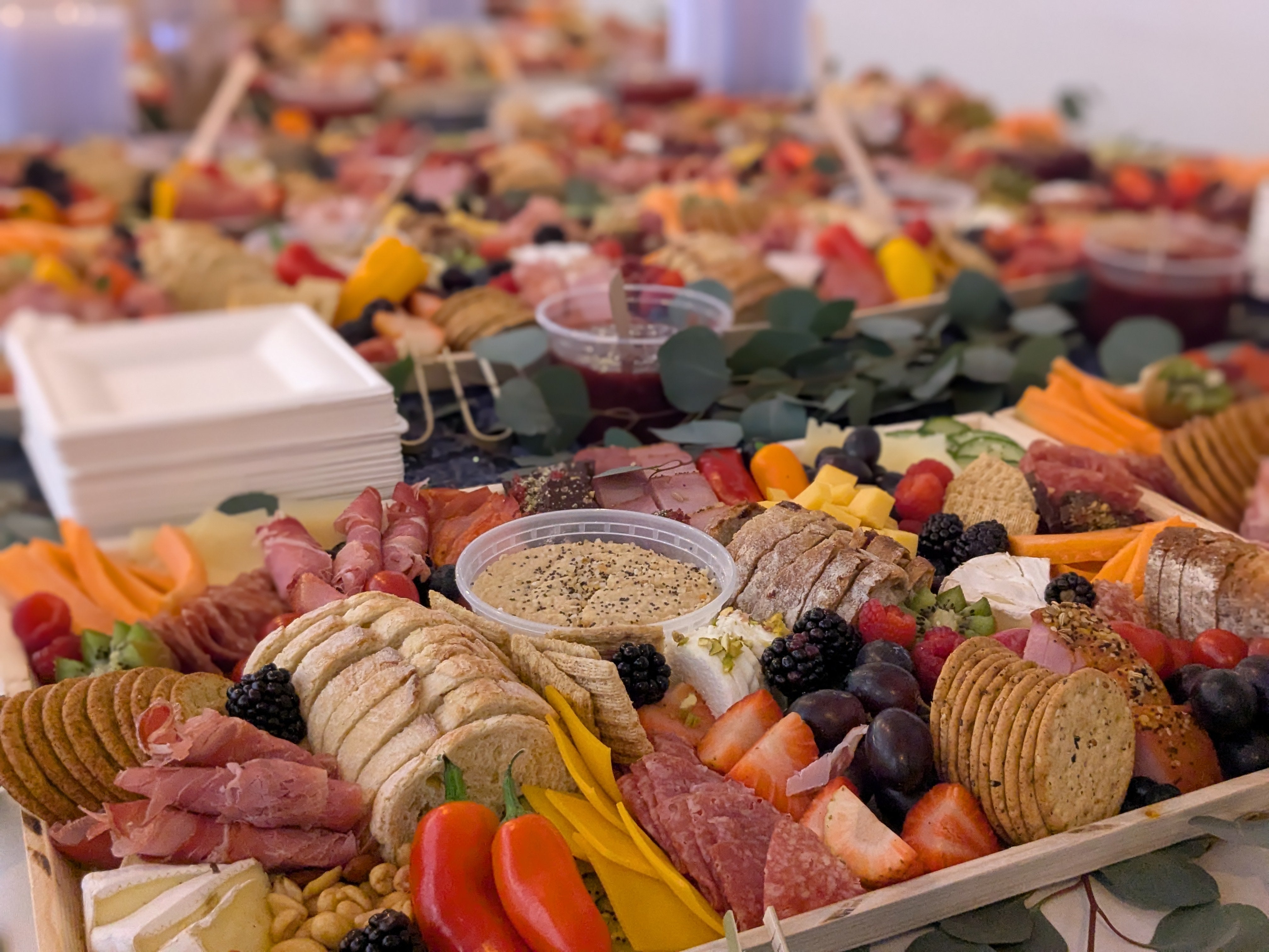 Charcuterie boards placed on a large table.  Displayed are meats, cheeses, chips, crackers, fruit and veggie options.  