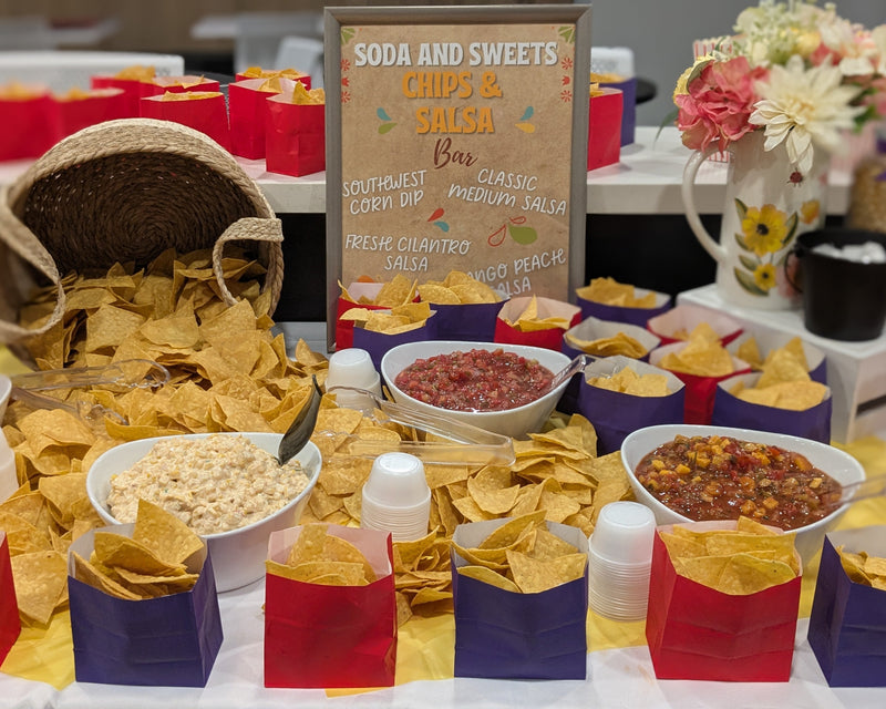 Snack bar with chips, salsa, and other items on a table with a sign indicating 'Soda and Sweets' and 'Chips & Salsa'.