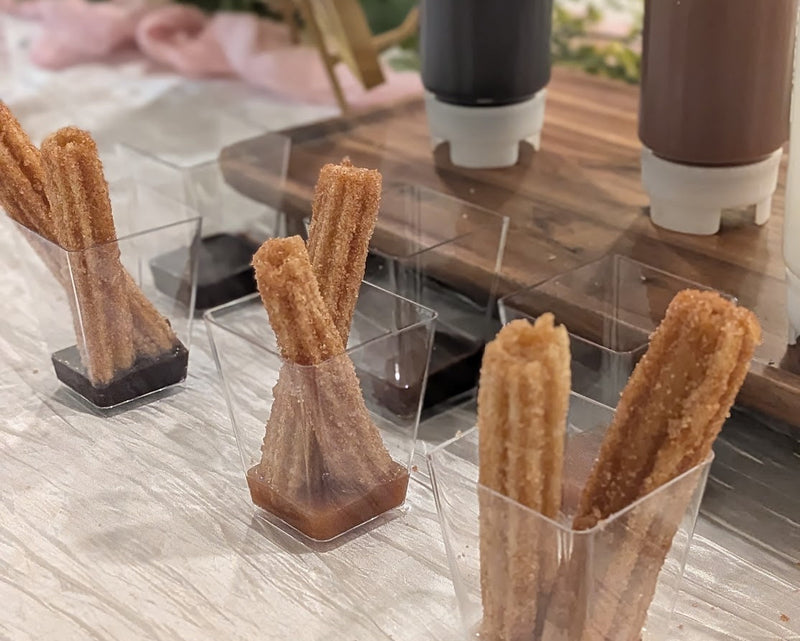 Small clear cups containing two churros in each with chocolate, carmel, or icing in the bottom to dip from.  