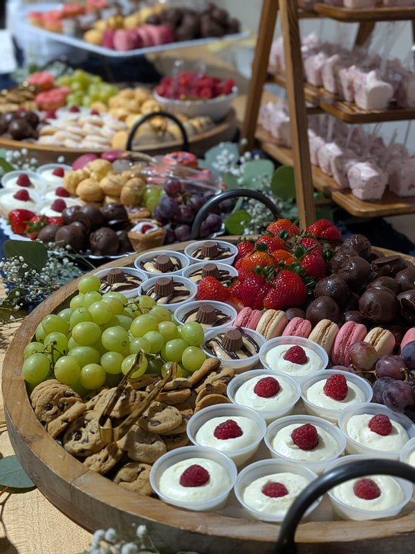 Table with multiple large trays of desserts including cookies, dessert cups, oreo balls, raspberry mousse, macaroons, and fresh in season fruit. 