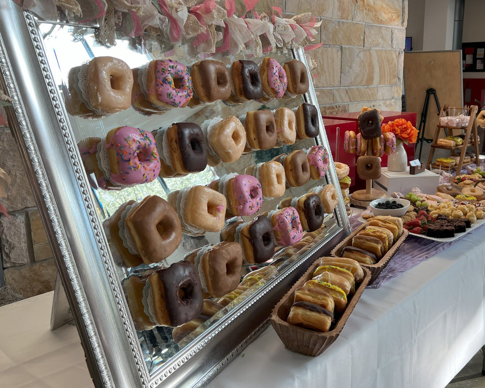 Donut display with various flavors on a table in a casual setting.  The display includes 24 donuts on a mirror display. and additional donuts on the table and platters.  