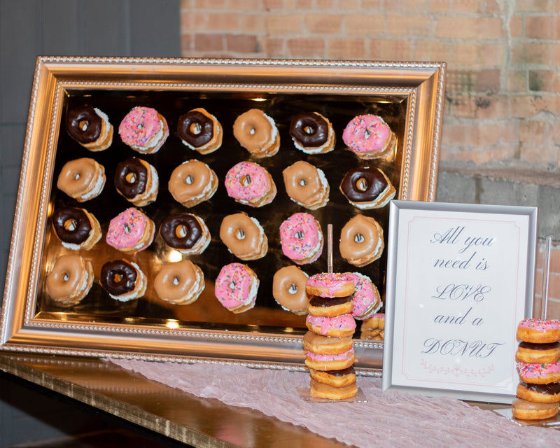 Donut display case with pink and chocolate donuts on a table against a brick wall.