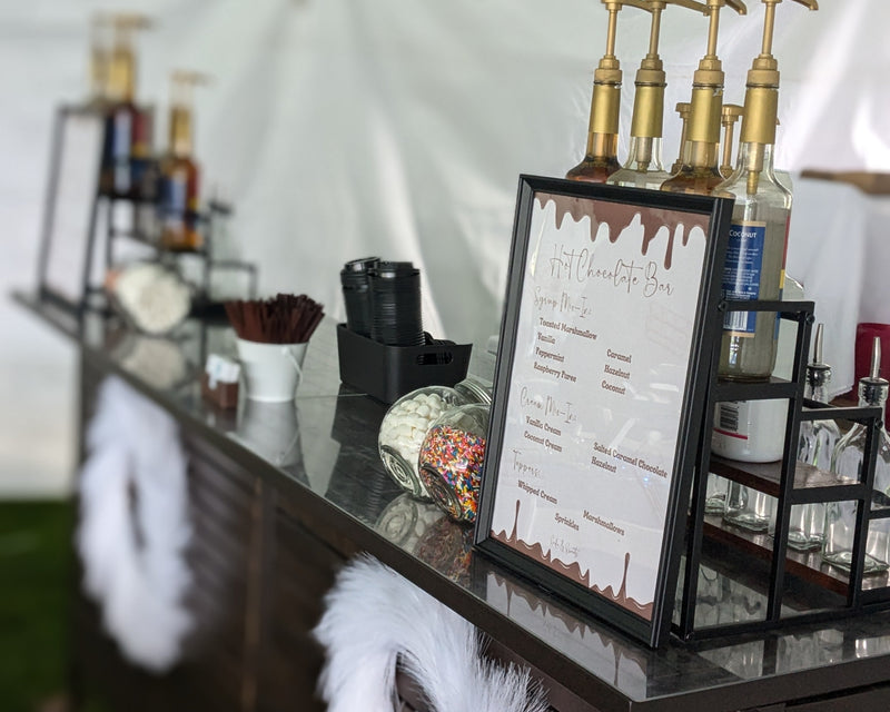 Hot chocolate bar setup with bottles and a menu board in an outdoor setting.  Large setup ready to serve hundreds of guests.
