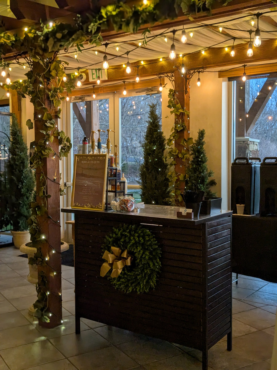 Wood colored serving bar configured for hot chocolate in front of a festive background of a trellace and lights.  