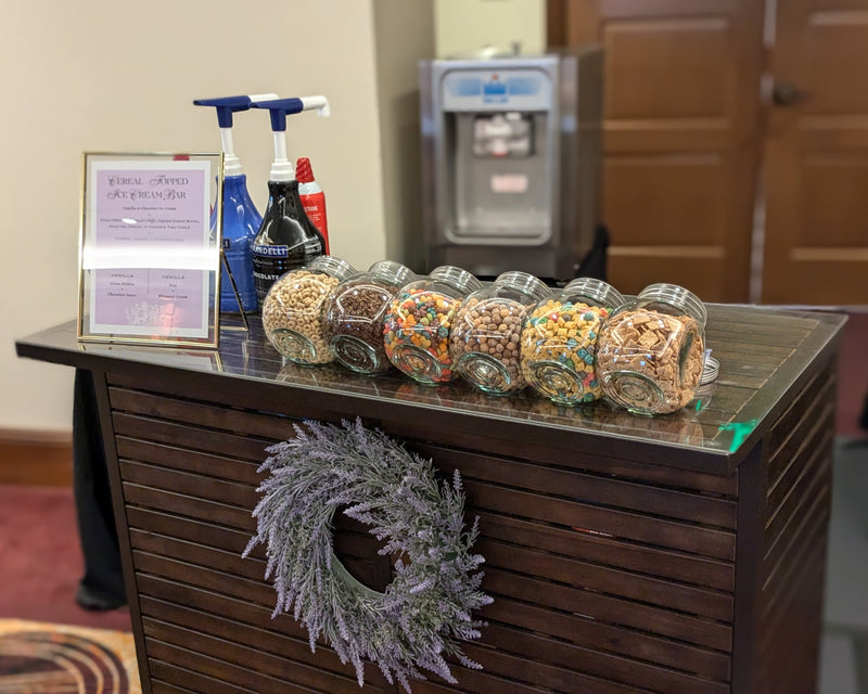 Ice cream sundae bar with decorative display with jars of ice cream toppings on a wooden stand in a room setting.