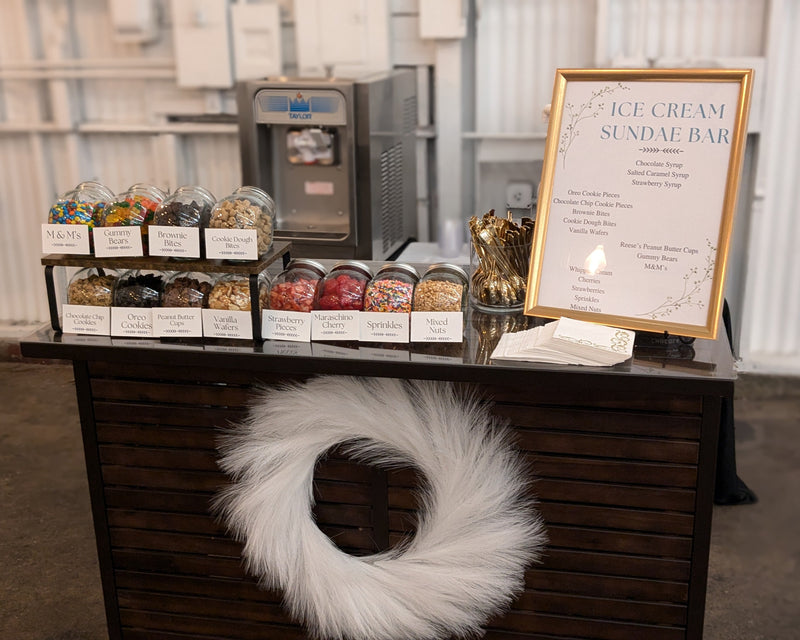 Ice cream sundae bar setup with ingredients and a sign on a wooden counter.