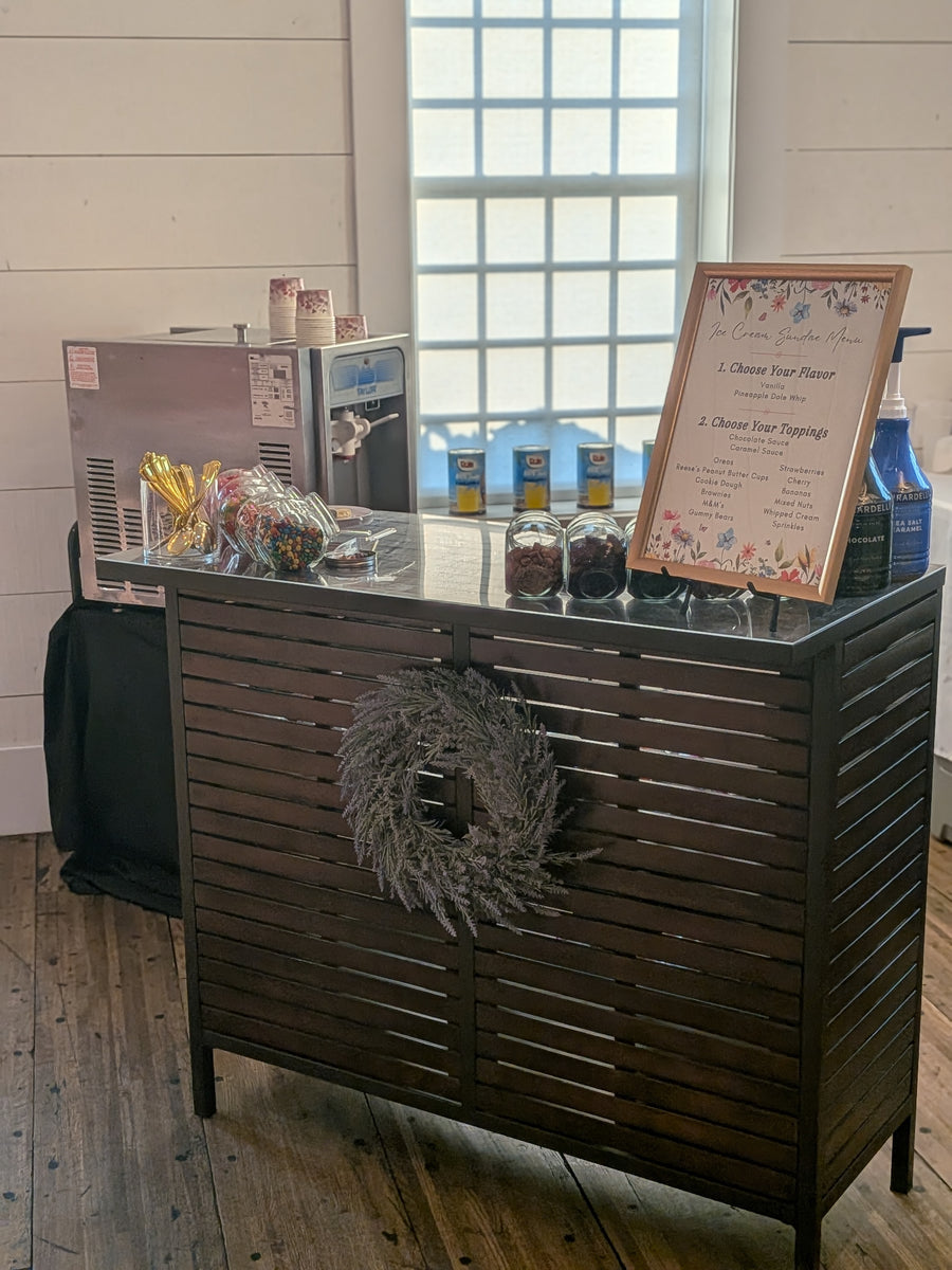 Ice cream sundae bar serving station, soft serve machine in the background.  Ice cream toppings and syrups are on the serving bar.  