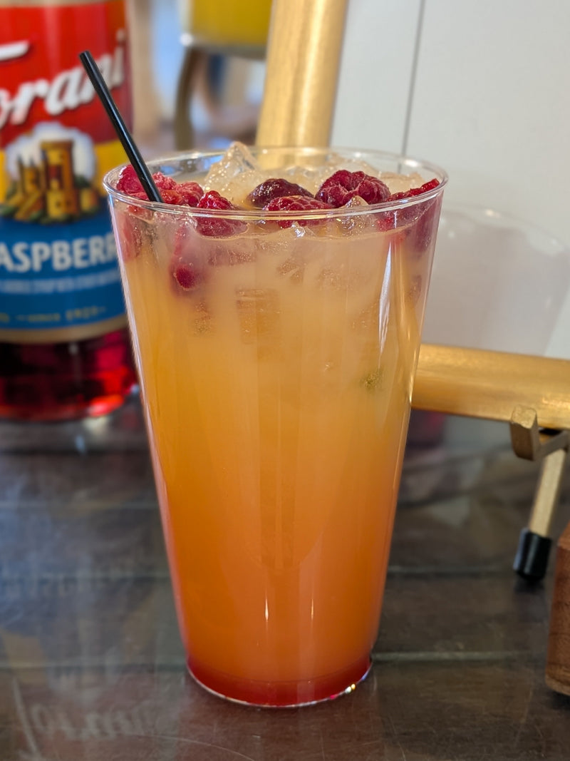 Orange drink non-alcoholic mocktail with raspberries in a glass with a straw, next to a bottle of Torani Raspberry syrup.