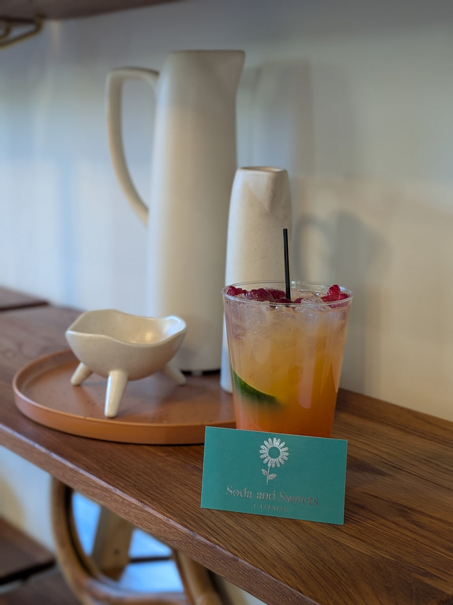 Colorful cocktail with ice and a straw on a wooden table, accompanied by a pitcher and small bowl. A card with 'Soda and Sweets Catering' is visible.