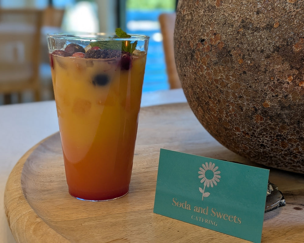 Fruity non-alcoholic mocktail blend of fresh juices, fruit in season, and garnishes on a table by a large rock vase.  A card showing "Soda and Sweets CAtering? is adjacent to the drink in a clear glass.  