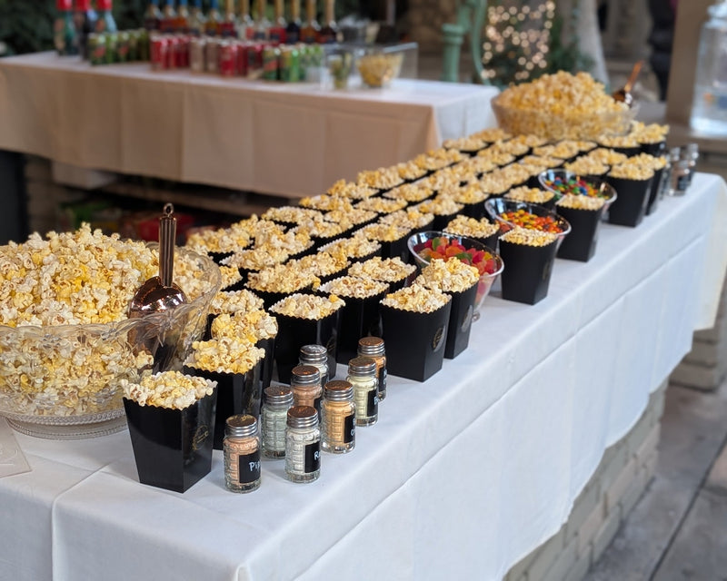 Freshly popped popcorn in boxes on a table,  toppings and flavor salts nearby.  