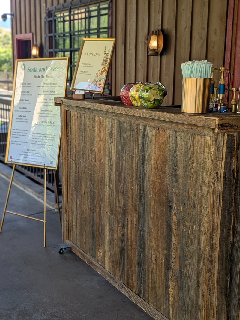 Wooden soda bar with a high countertop displaying lemons, limes, strawberries, and syrups to mix in with a wide range of popular soda flavors and lemonades.   Straws and the menu are on the edges of the photo.  