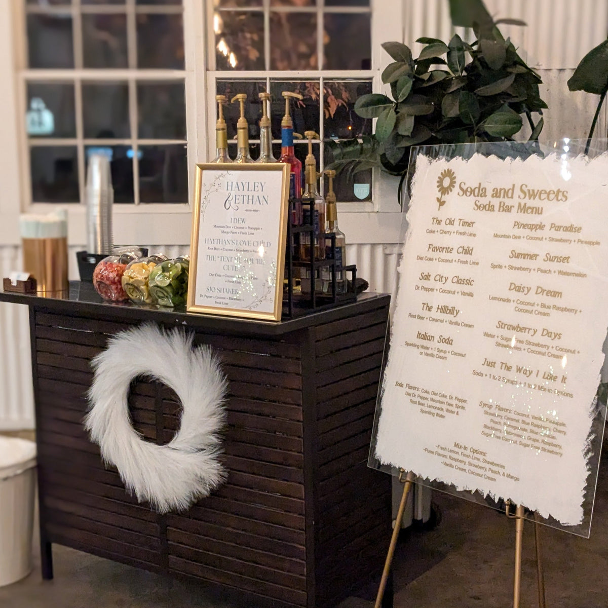 Soda, Soda Bar, Flavored Soda, Syrups, Menu, Lemons, Limes, Strawberries.  Brown serving bar with a white wreath, a large menu on an easel. 