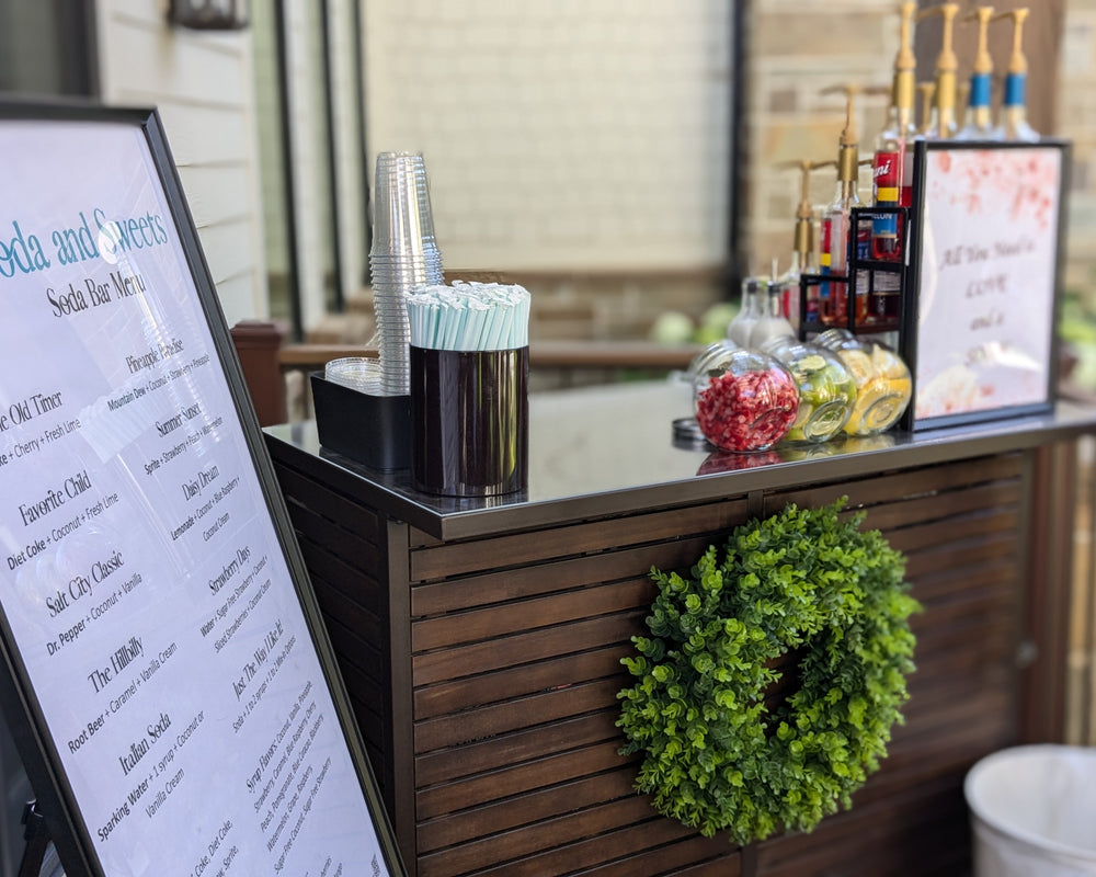 Outdoor bar setup with drink menus, cups, and decorative wreath.  Soda bar, soda options, flavored sodas.