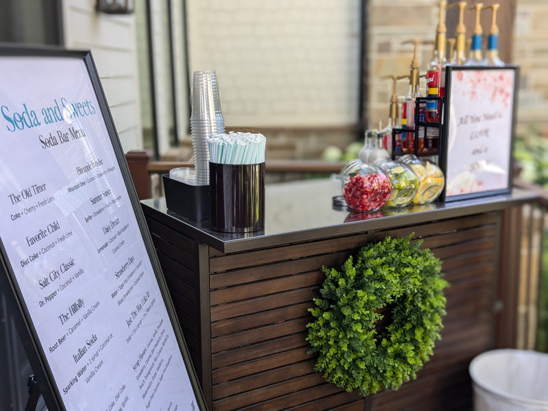 Soda bar.  Outdoor bar setup with flavored soda drink menus, cups, and decorative wreath.