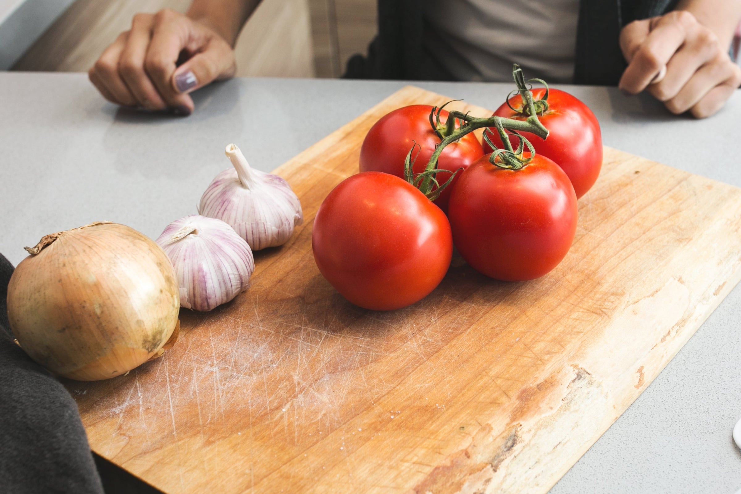 Tomatoes on a wooden cutting board with garlic and an onion, on a kitchen counter.